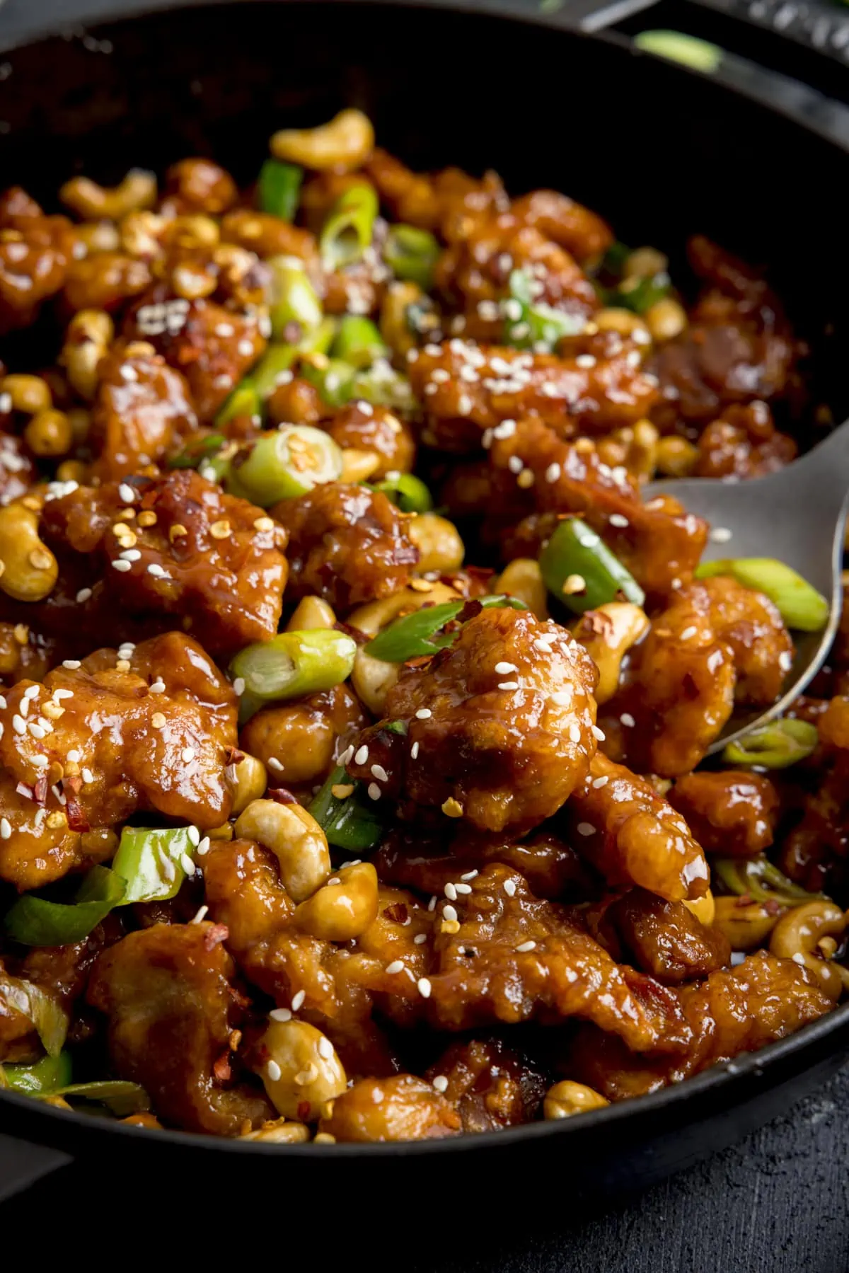 Close up of crispy chicken with cashews in a sticky sauce with spring onions in a black pan. A spoonful of chicken is being taken from the pan.