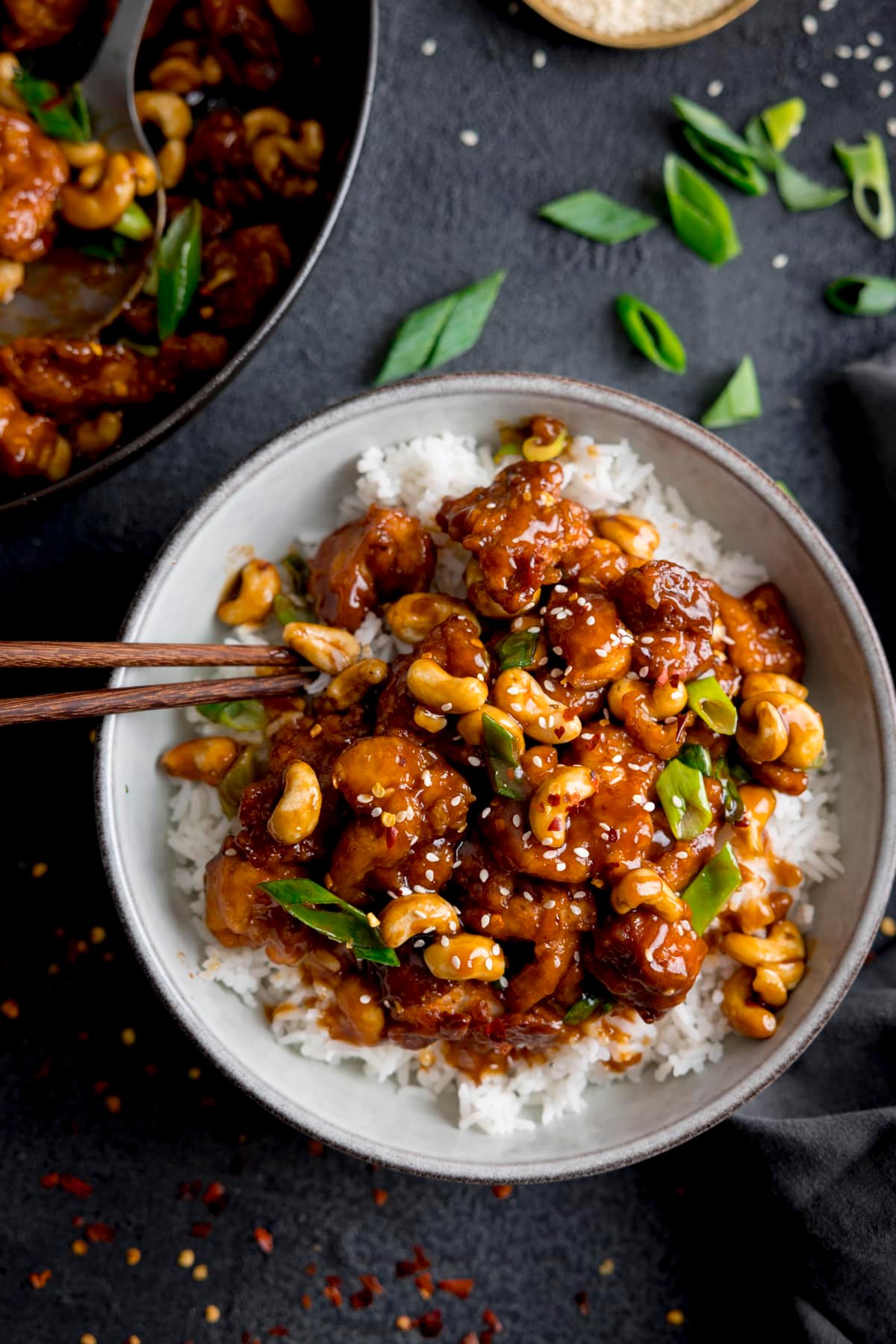 Overhead of sticky cashew chicken with rice in a white bowl on a dark background. There are chopsticks sticking out of the bowl. The pan of cashew chicken is in shot and there are chopped spring onions sprinkled around.