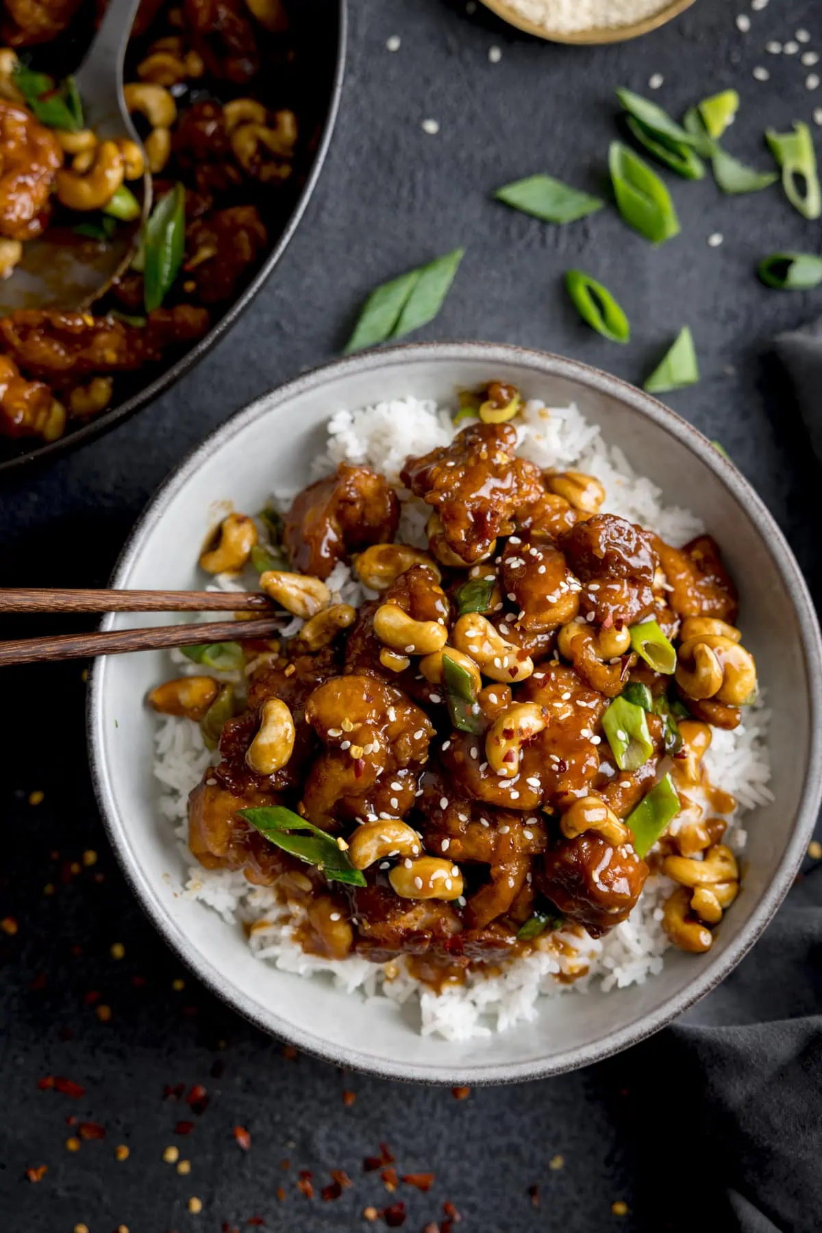 Overhead of sticky cashew chicken with rice in a white bowl on a dark background. There are chopsticks sticking out of the bowl. The pan of cashew chicken is in shot and there are chopped spring onions sprinkled around.