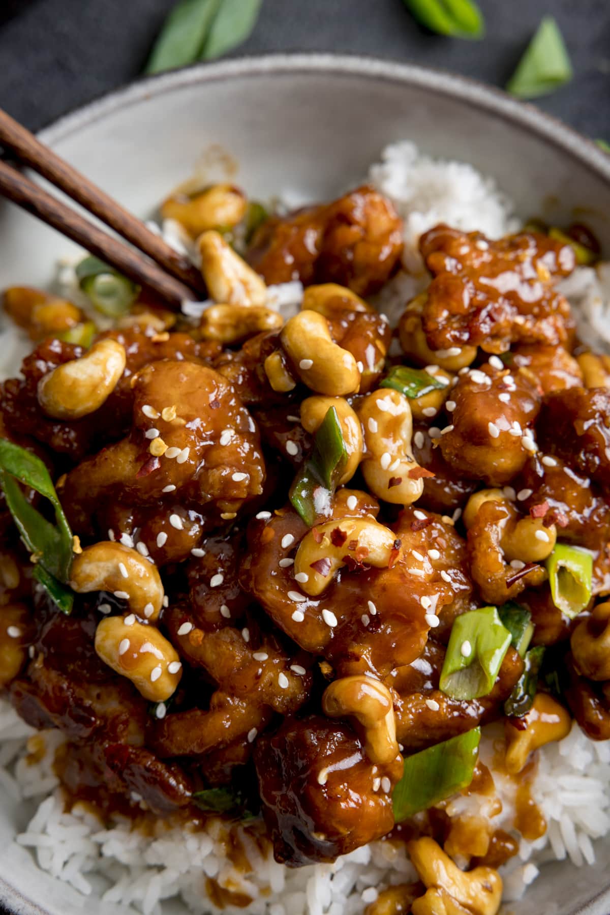 Close up of crispy chicken with cashews in a sticky sauce with spring onions in a bowl with rice. There are sesame seeds sprinkled on top and a pair of chopsticks sticking out of the dish