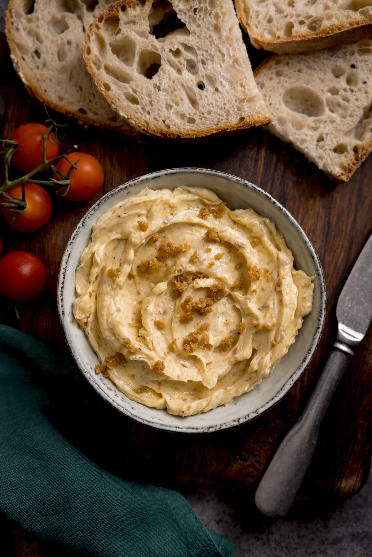 Overhead image of chicken skin butter in a grey bowl on a wooden board. There are slices of bread, some vine cherry tomatoes, a knife and a green napkin around the bowl.
