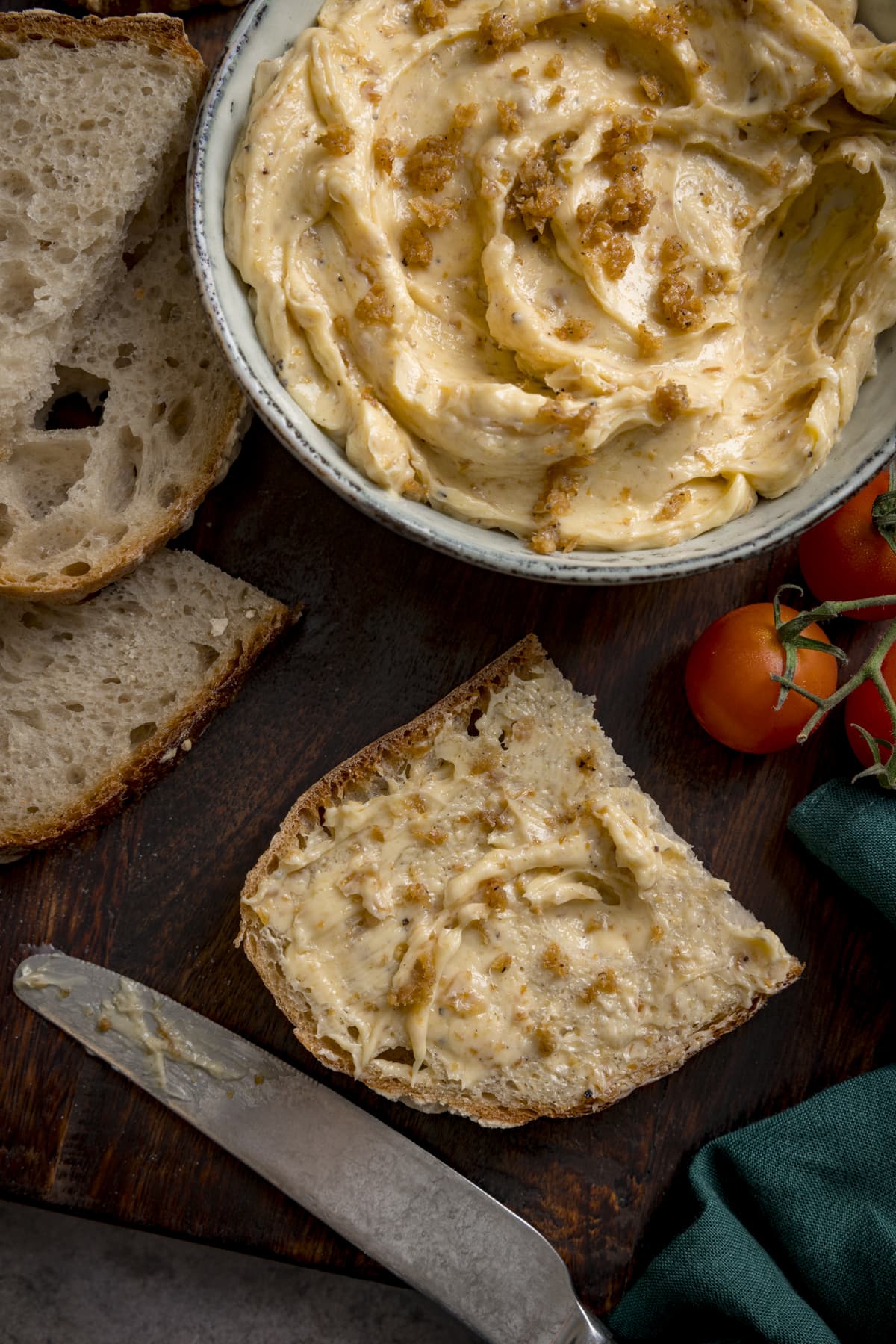A bowl of chicken skin butter and a piece of bread with the butter spread on it. There is also a knife, a couple of cherry tomatoes and sliced of un-buttered bread scattered around.
