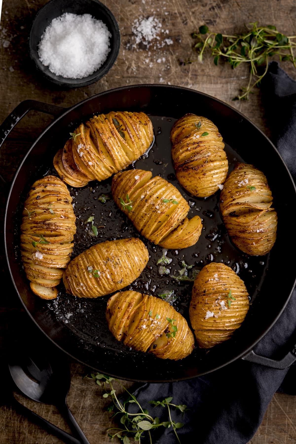 Top view of Hasselback potatoes in a dark cast iron skillet, garnished with fresh thyme leaves and Maldon salt.  The pan is on a wooden table and around the pan is a small bowl of Maldon salt and some thyme leaves.