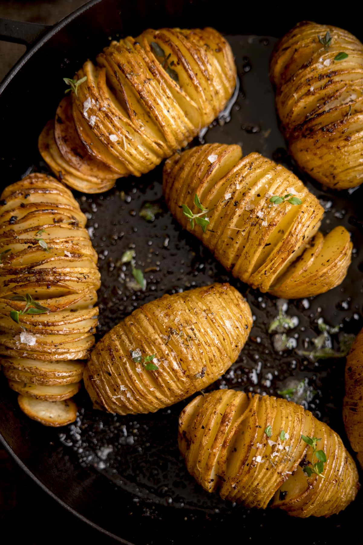 Top view of Hasselback potatoes in a dark cast iron skillet, garnished with fresh thyme leaves and Maldon salt