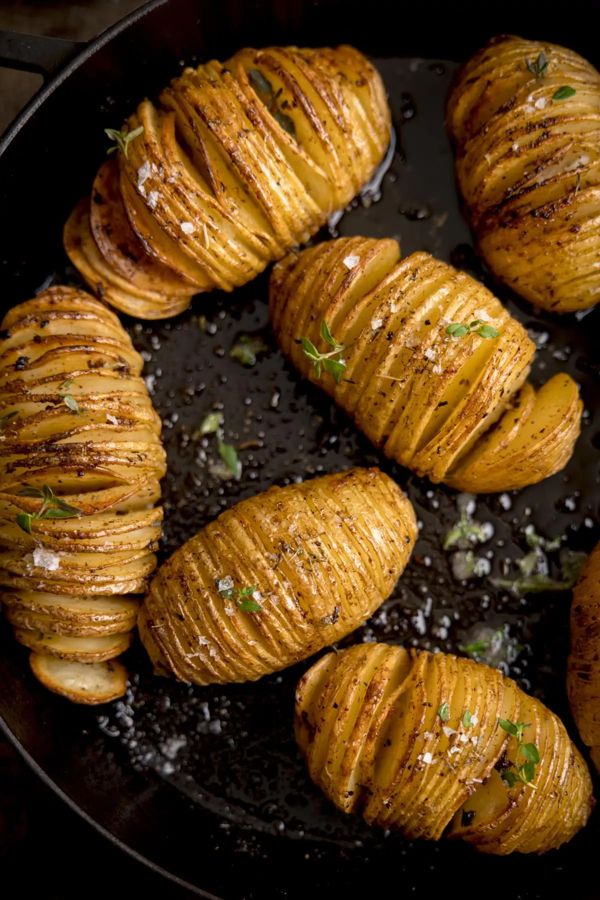 Overhead image of hasselback potatoes in a dark cast iron pan, topped with fresh thyme leaves and Maldon salt