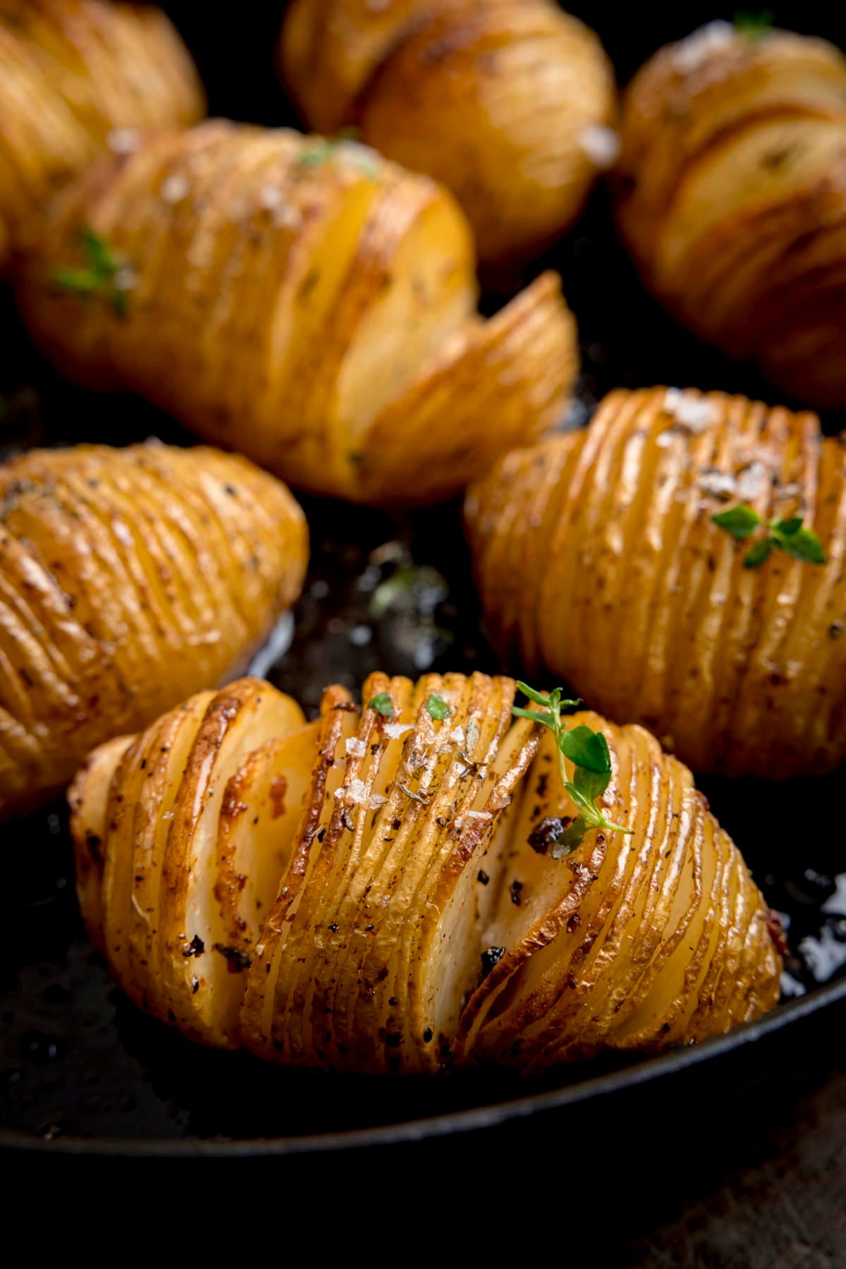 Close-up of Hasselback potatoes in a dark cast iron skillet, garnished with fresh thyme leaves and Maldon salt
