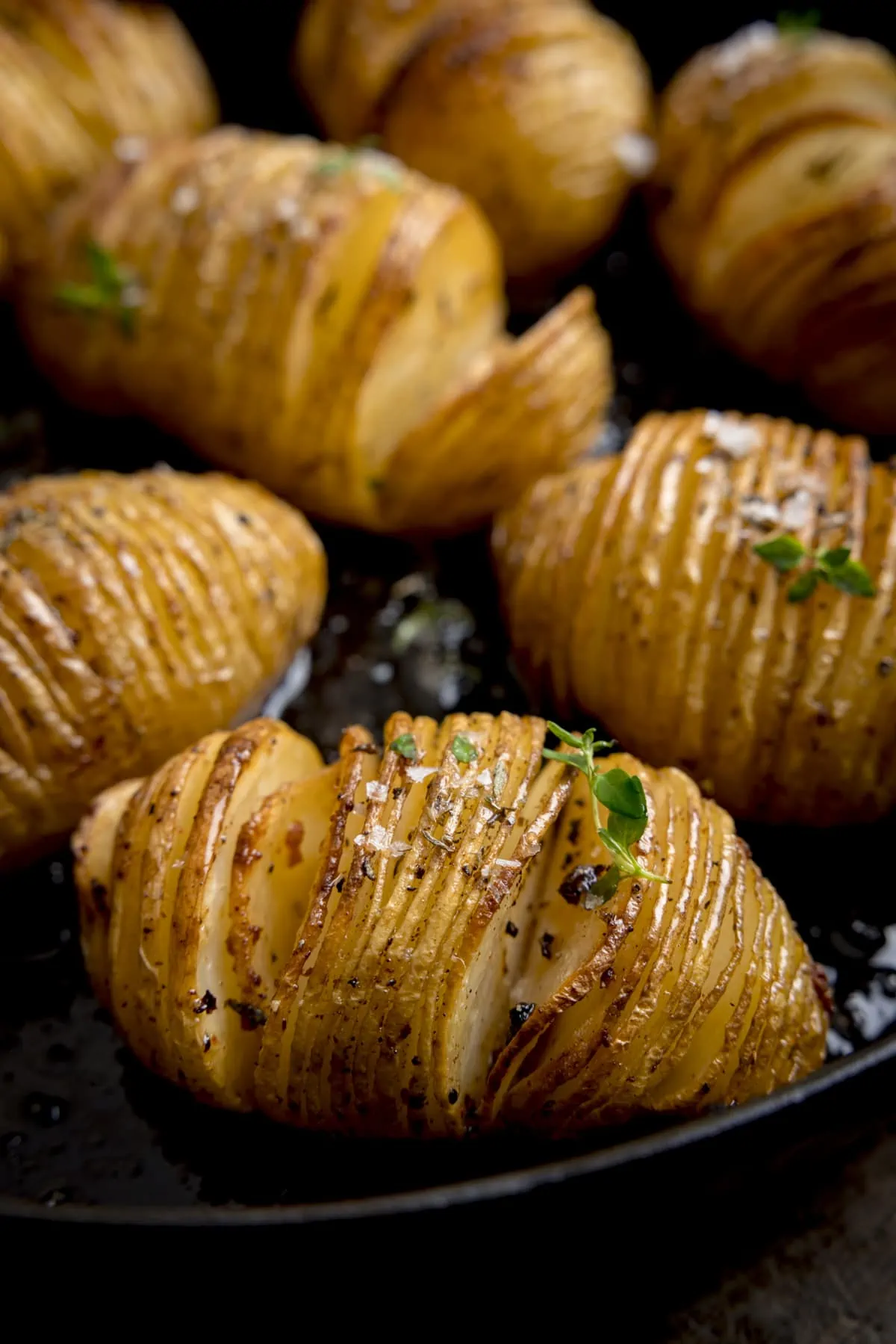 Close up image of hasselback potatoes in a dark cast iron pan, topped with fresh thyme leaves and Maldon salt