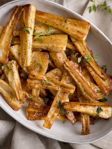 Overhead image of Honey roast parsnips, topped with thyme leaves in a white oval bowl. The bowl is on a beige napkin.