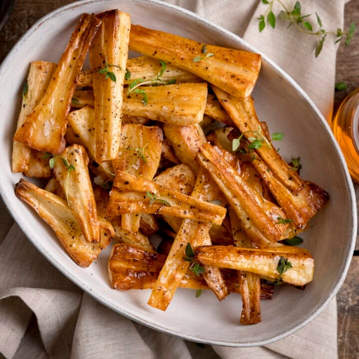 Overhead image of Honey roast parsnips, topped with thyme leaves in a white oval bowl. The bowl is on a beige napkin.