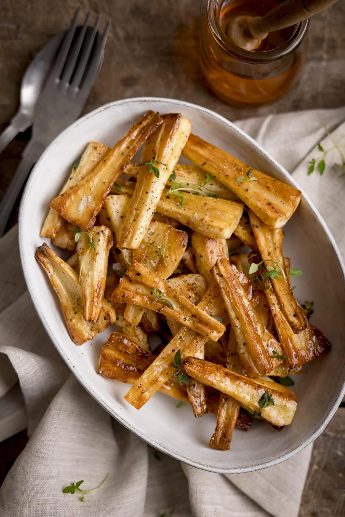 Overhead image of Honey roast parsnips, topped with thyme leaves in a white oval bowl. The bowl is on a beige napkin and there is a glass jar of honey next to it.