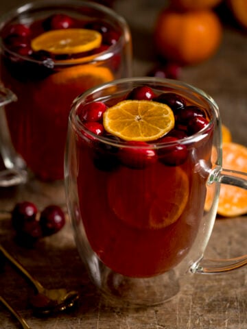 Square image of 2 glass mugs filled with mulled cider. There are clementine slices and cranberries in the cider and the mugs are on a wooden table.