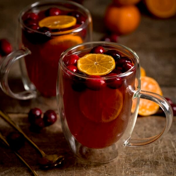 Square image of 2 glass mugs filled with mulled cider. There are clementine slices and cranberries in the cider and the mugs are on a wooden table.