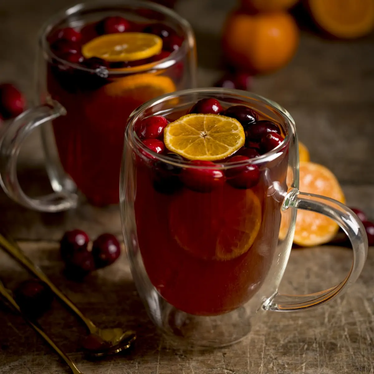 Square image of 2 glass mugs filled with mulled cider. There are clementine slices and cranberries in the cider and the mugs are on a wooden table.