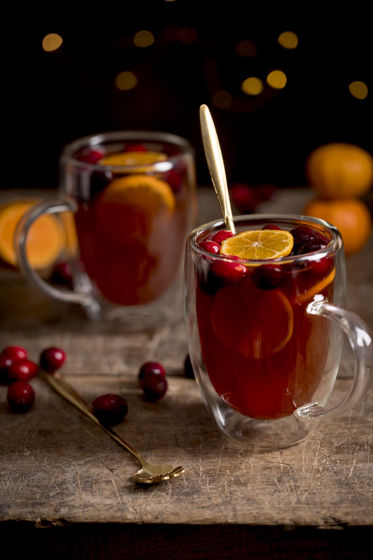 2 glass mugs filled with mulled cider. There are clementine slices and cranberries in the cider and the mugs are on a wooden table. The front mug has a gold teaspoon in it.