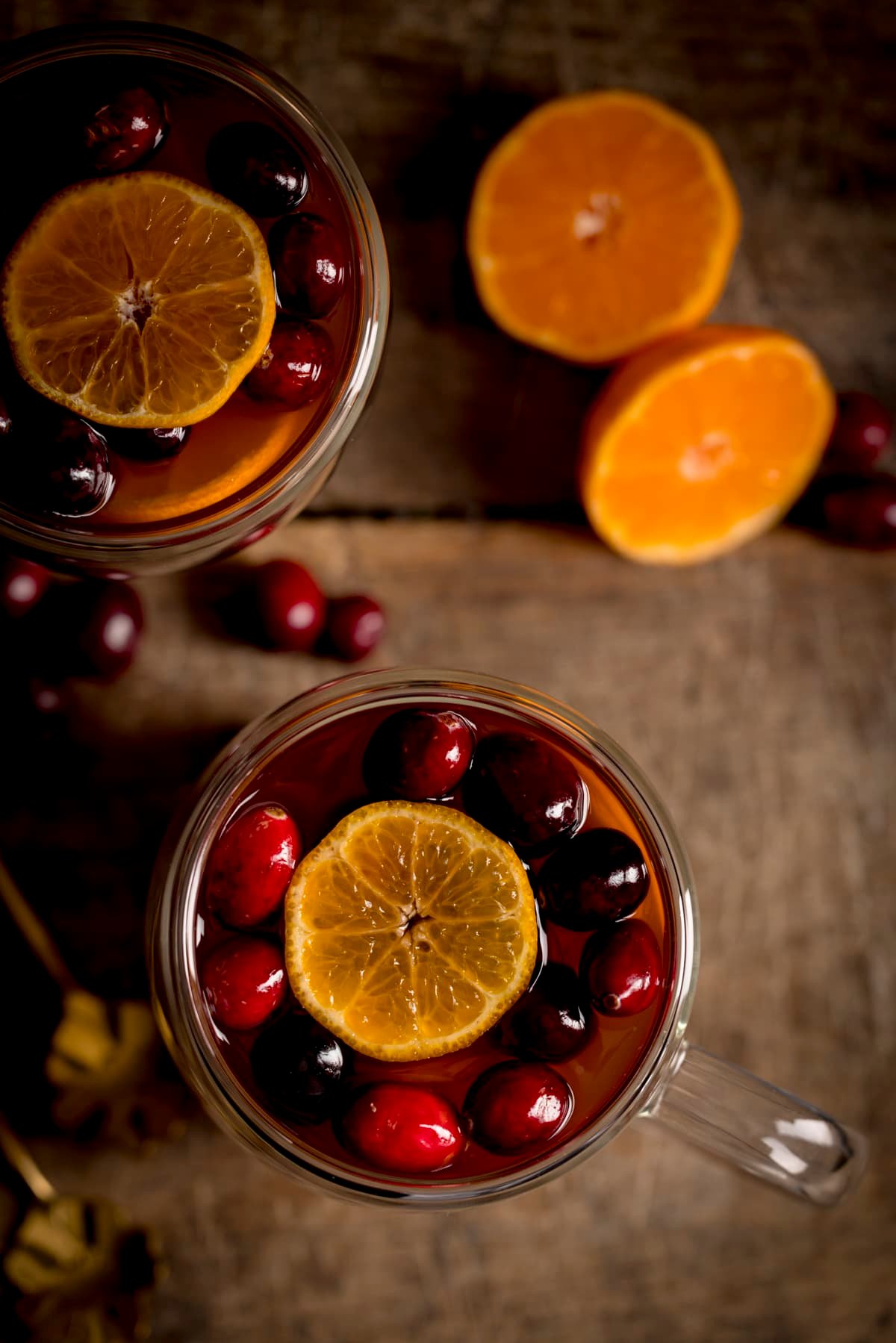 Overhead image of two glass mugs filled with mulled cider, cranberries and clementine slices on a wooden table. There is a clementine sliced in half next to the mugs.