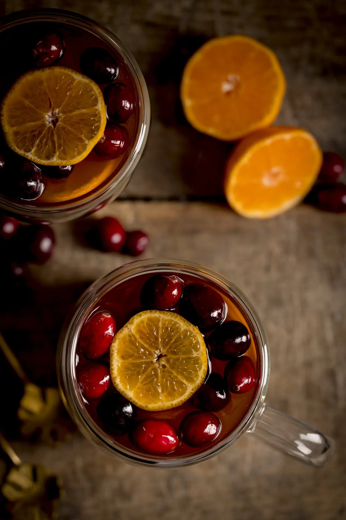 Overhead image of two glass mugs filled with mulled cider, cranberries and clementine slices on a wooden table. There is a clementine sliced in half next to the mugs.