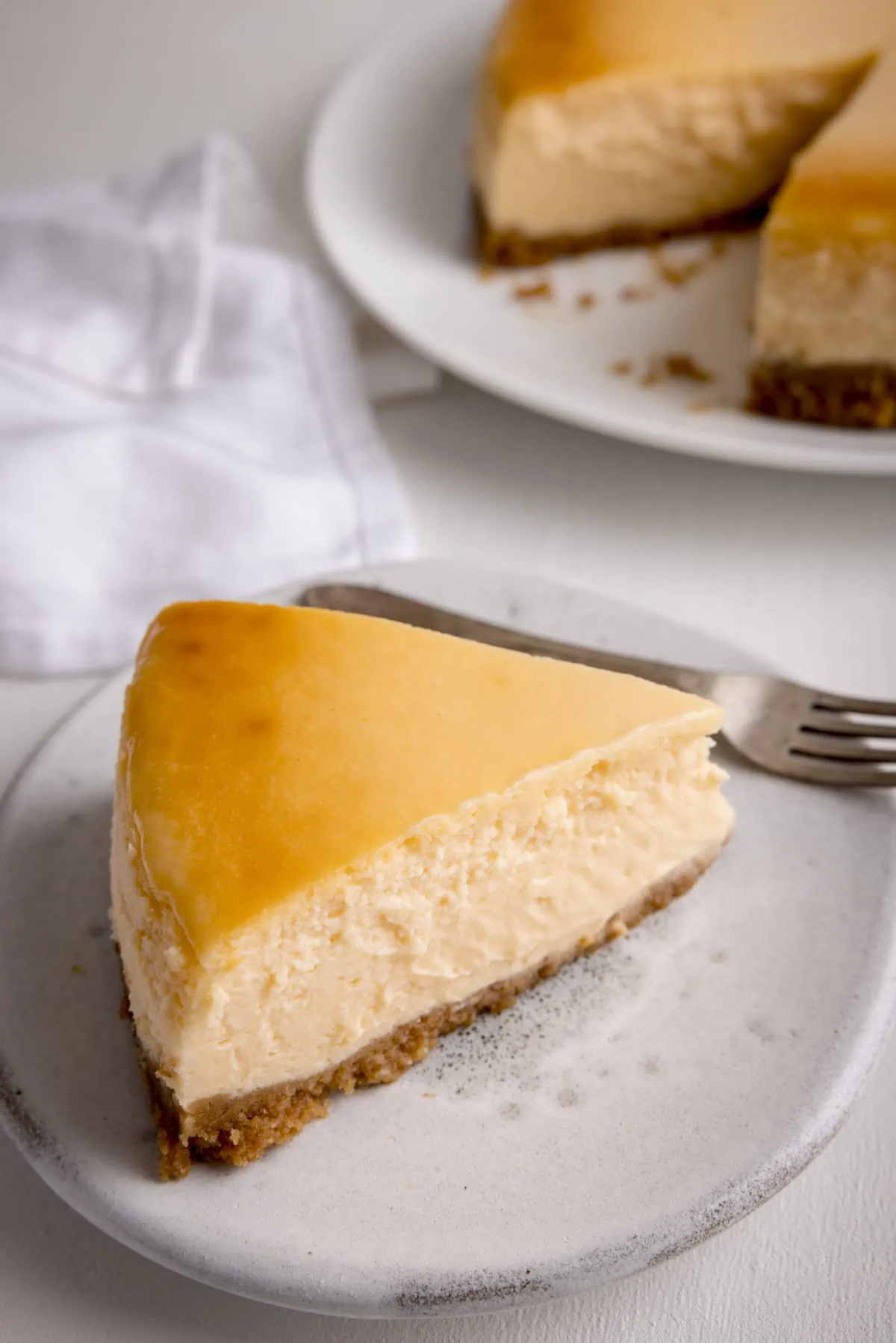 A slice of New York Cheesecake on a white plate on a white background. The rest of the cheesecake is just in shot at the top of the image.