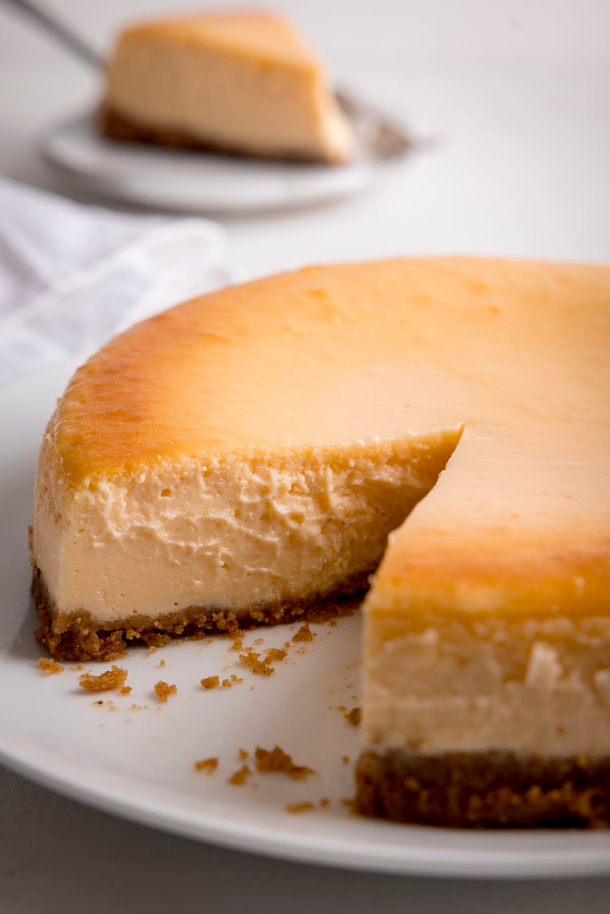 Plain New York cheesecake on a light background with one slice taken out.  The slice lies on a plate in the background.