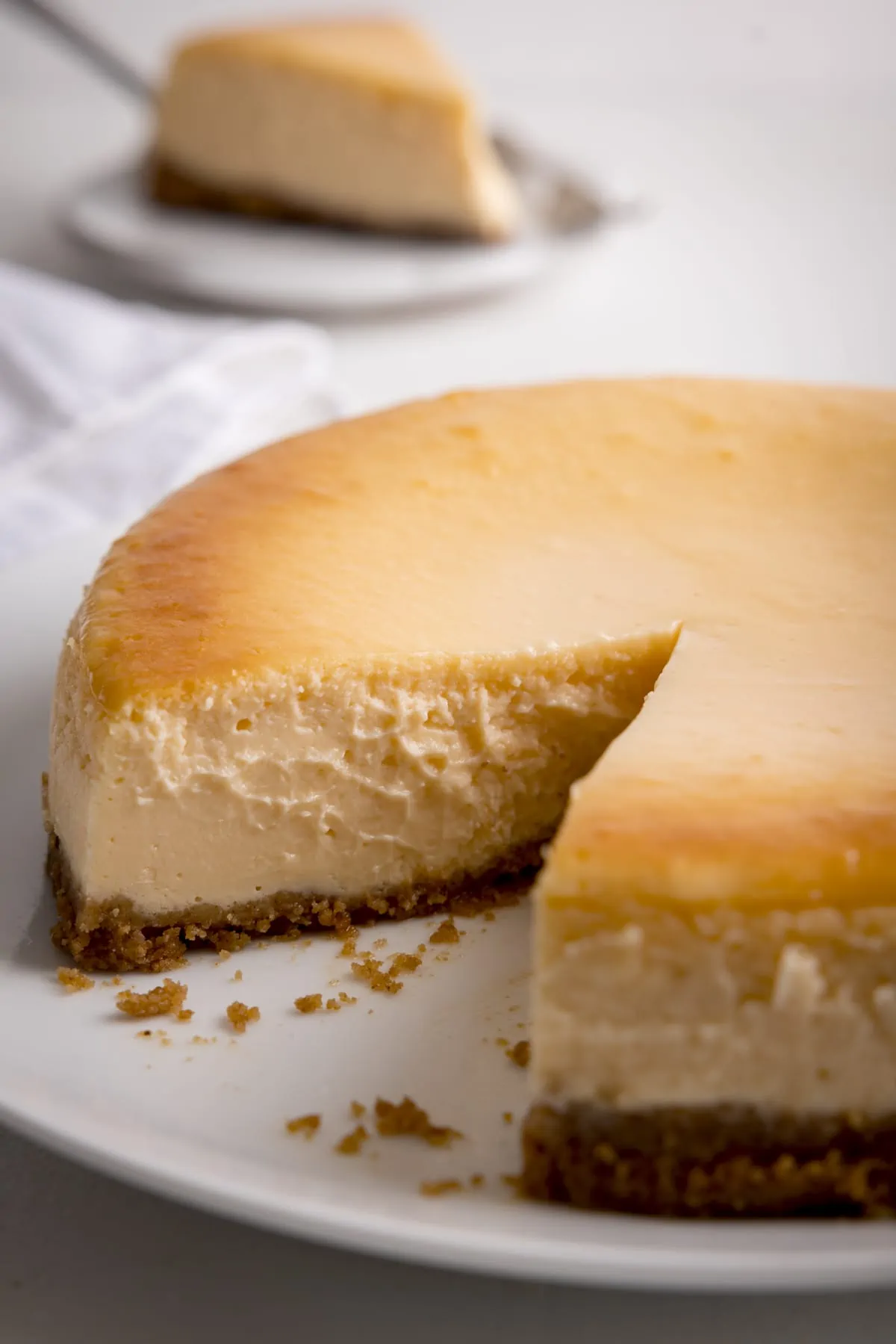 Plain New York Cheesecake on a light background with a slice taken out. The slice is on a plate in the background.