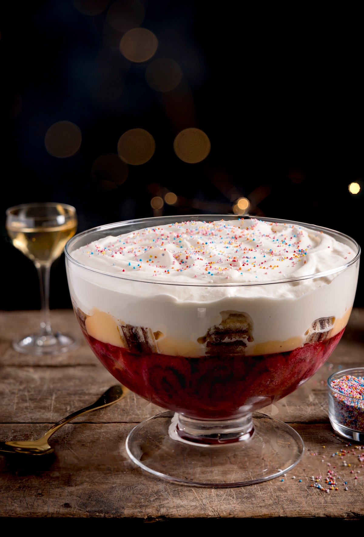 Sherry trifle in a large glass dish on a wooden table against a dark background. There is a glass of sherry and a bowl of sprinkles also in shot.