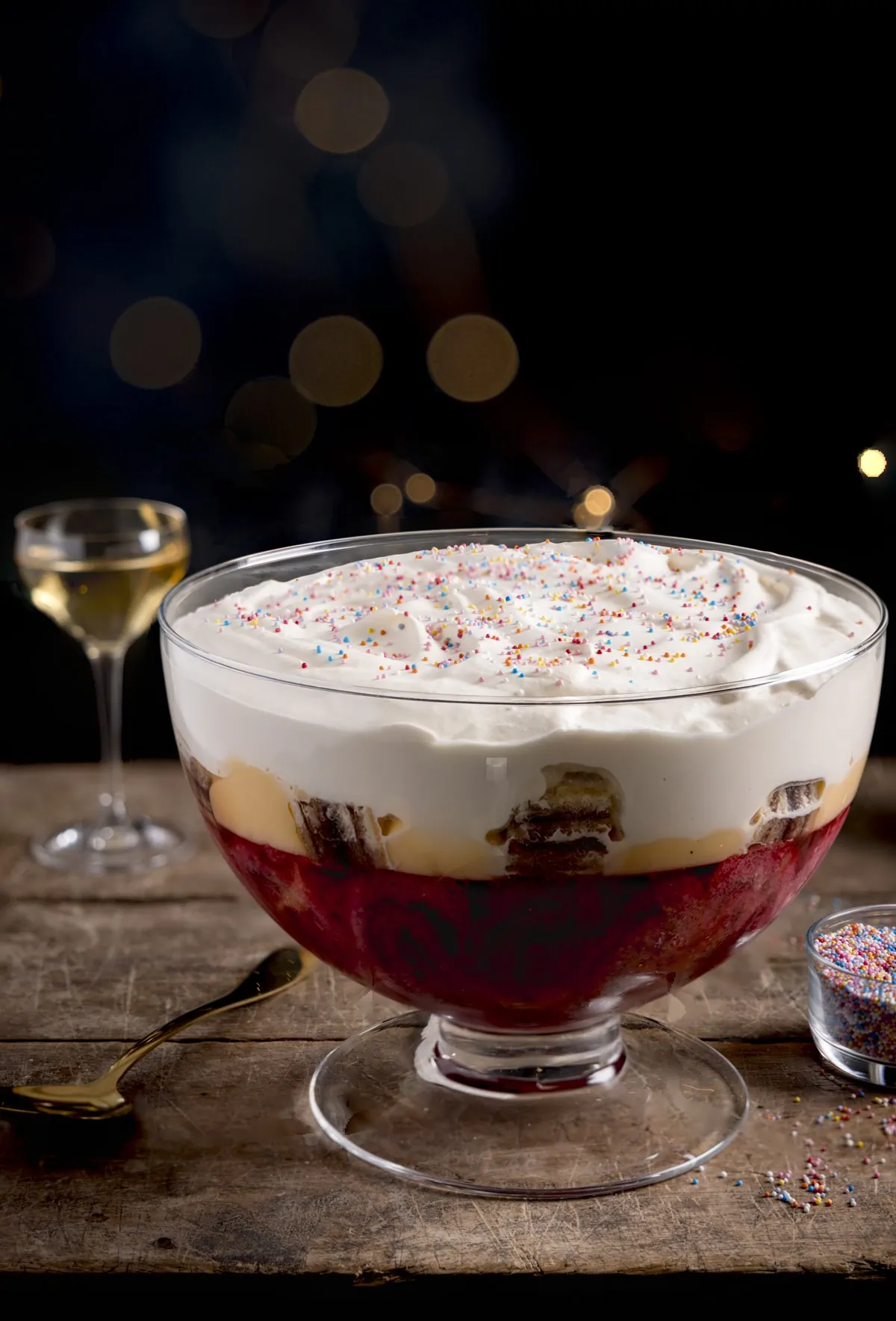 Sherry trifle in a large glass dish on a wooden table against a dark background. There is a glass of sherry and a bowl of sprinkles also in shot.