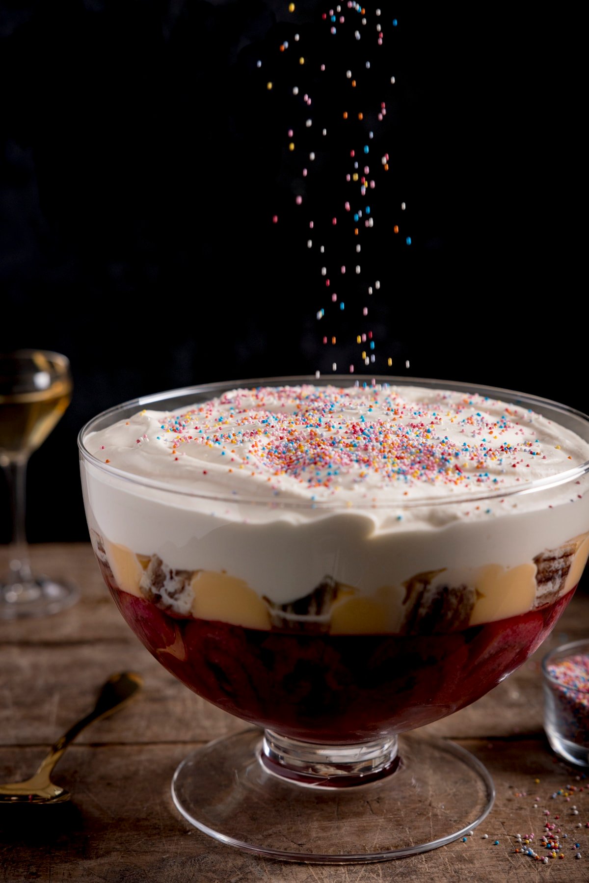 Rainbow sprinkles being sprinkled onto a large bowl of sherry trifle. The trifle is on a wooden table, against a dark background.