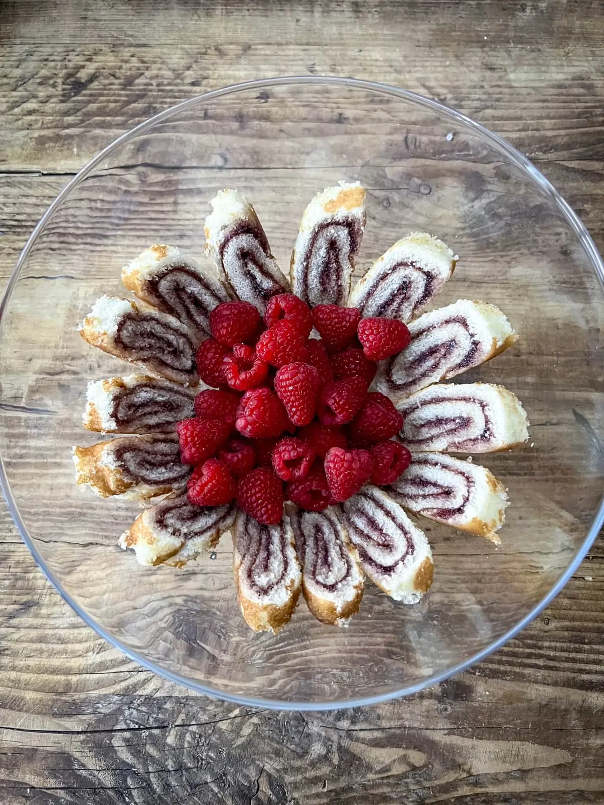Overhead shot of glass trifle bowl with slices of jam swiss roll and fresh raspberries in. The bowl is on a wooden table.