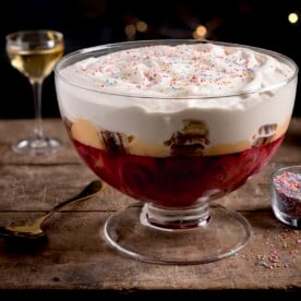 Square image of sherry trifle in a large glass dish on a wooden table against a dark background. There is a glass of sherry and a bowl of sprinkles also in shot.