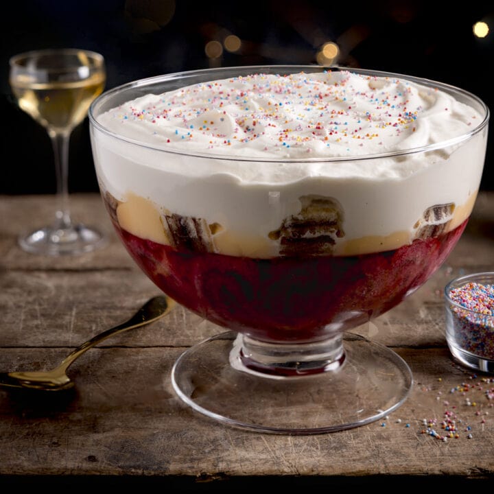 Square image of sherry trifle in a large glass dish on a wooden table against a dark background. There is a glass of sherry and a bowl of sprinkles also in shot.