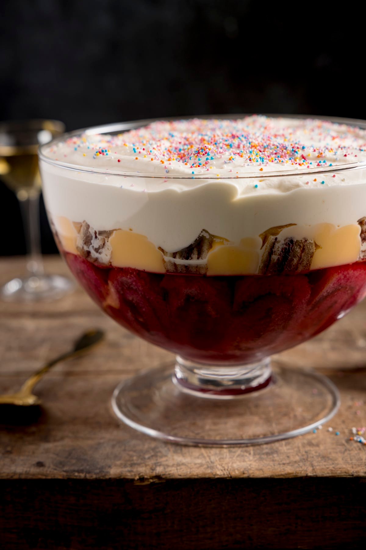 Side image of a large glass bowl of sherry trifle on a wooden table against a dark background.