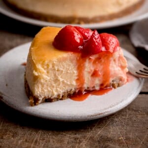 صورة جانبية مربعة لشريحة من كعكة الجبن بالفراولة على طبق أبيض على طاولة خشبية.  هناك شوكة حلوى والمزيد من كعكة الجبن في الخلفية ، فقط في لقطة.
