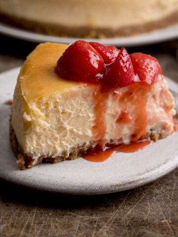 Square side-on image of a slice of strawberry cheesecake on a white plate on a wooden table. There is a dessert fork and more cheesecake in the background, only just in shot.