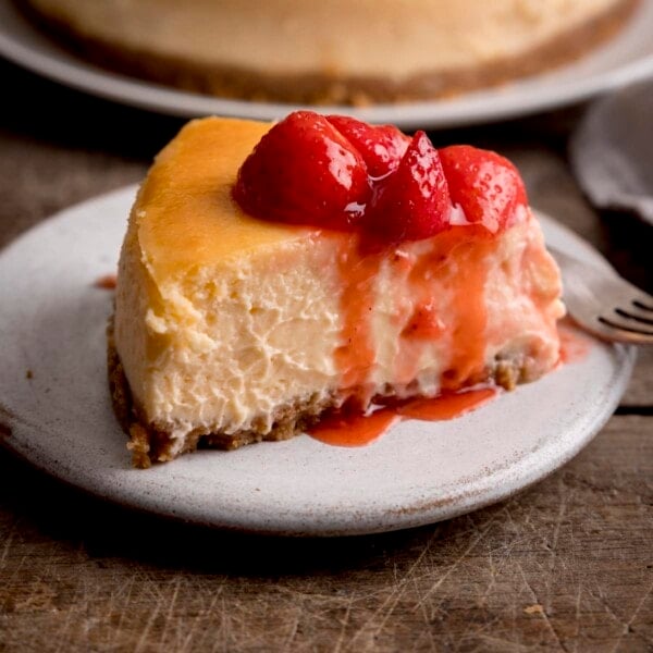 Square side-on image of a slice of strawberry cheesecake on a white plate on a wooden table. There is a dessert fork and more cheesecake in the background, only just in shot.