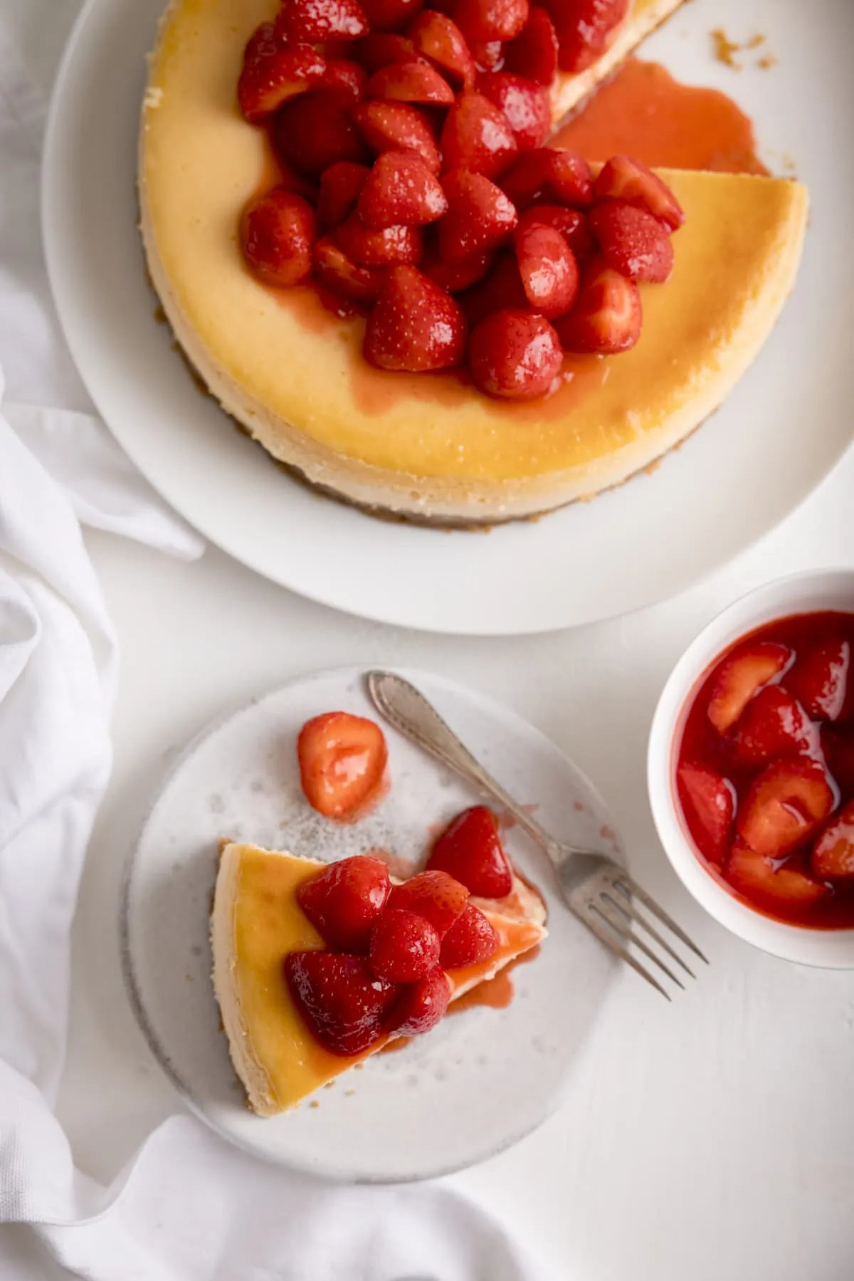Overhead shot of a slice of strawberry cheesecake on a white plate next to the rest of the cheesecake and a white bowl filled with strawberry compote. Everything is set on a white background.