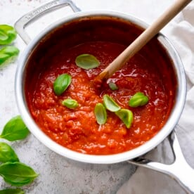 Square image of a silver pan filled with arrabbiata sauce on a light background. There is a wooden spoon and sprinkled fresh basil leaves in the sauce.