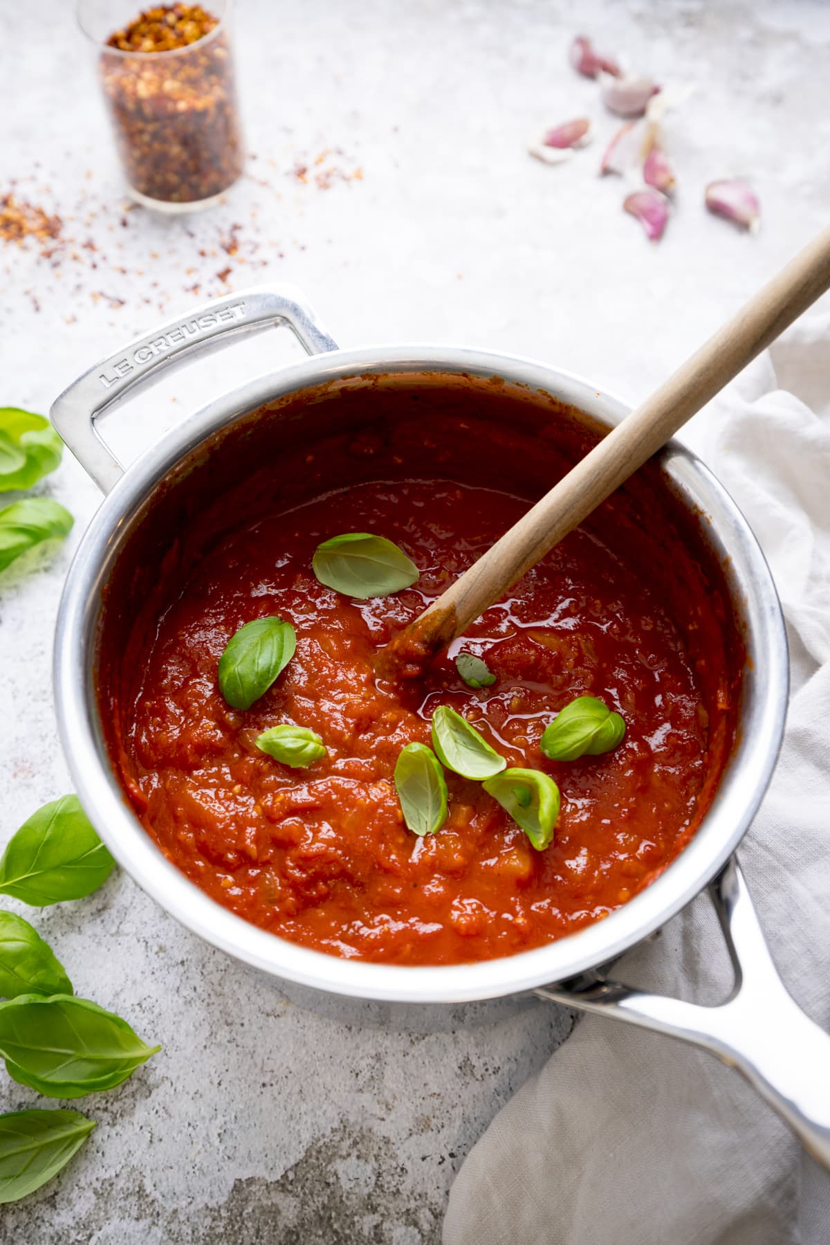 Overhead image of a silver pan filled with arrabbiata sauce on a light background. There is a wooden spoon and sprinkled fresh basil leaves in the sauce. There are basil leaves, garlic cloves and a jar of chilli flakes around the pan.