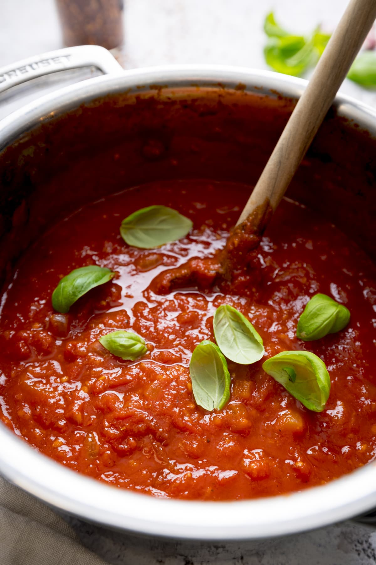Close up image of a silver pan filled with arrabbiata sauce on a light background. There is a wooden spoon and sprinkled fresh basil leaves in the sauce.