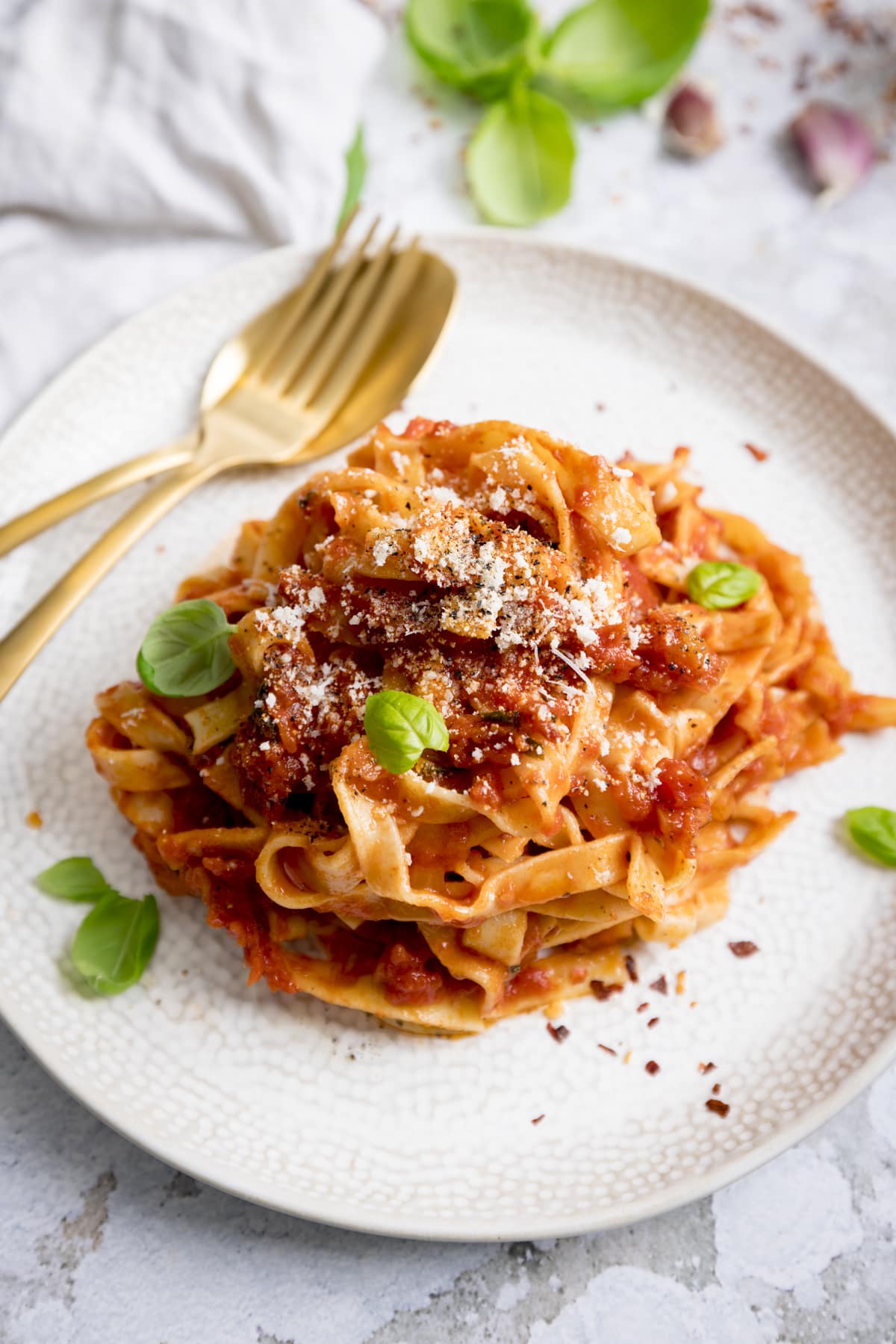 Tagliatelle in Arrabbiata sauce on a white plate on a light background. There is a gold fork and spoon on the plate and the pasta is sprinkled with parmesan, black pepper and basil leaves.