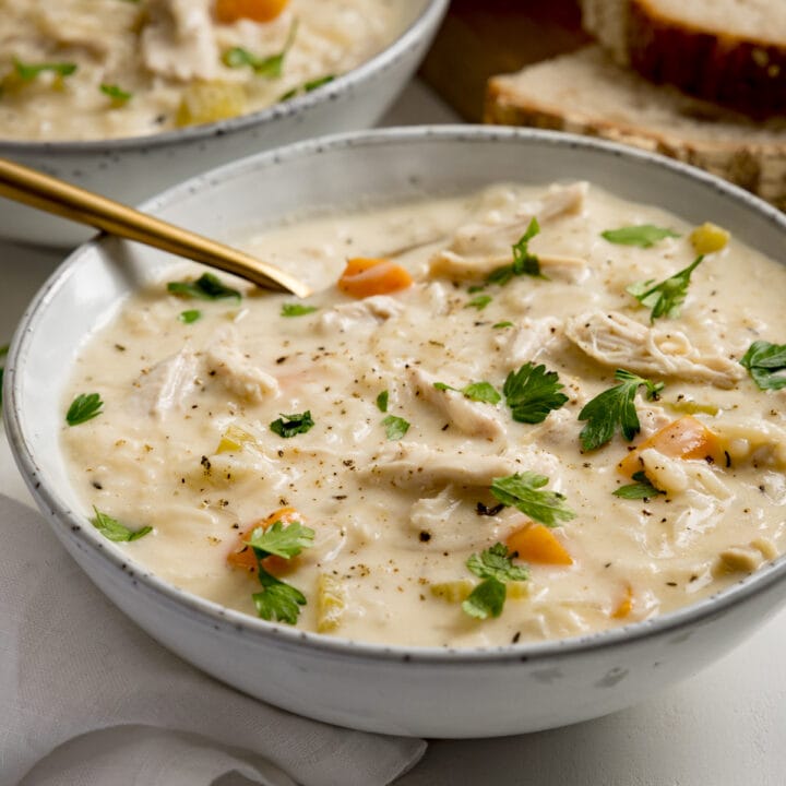 Square image of a bowl of chicken and rice soup on a light background. There is a gold spoon in the bowl. There is a further bowl and some bread slices in the background.