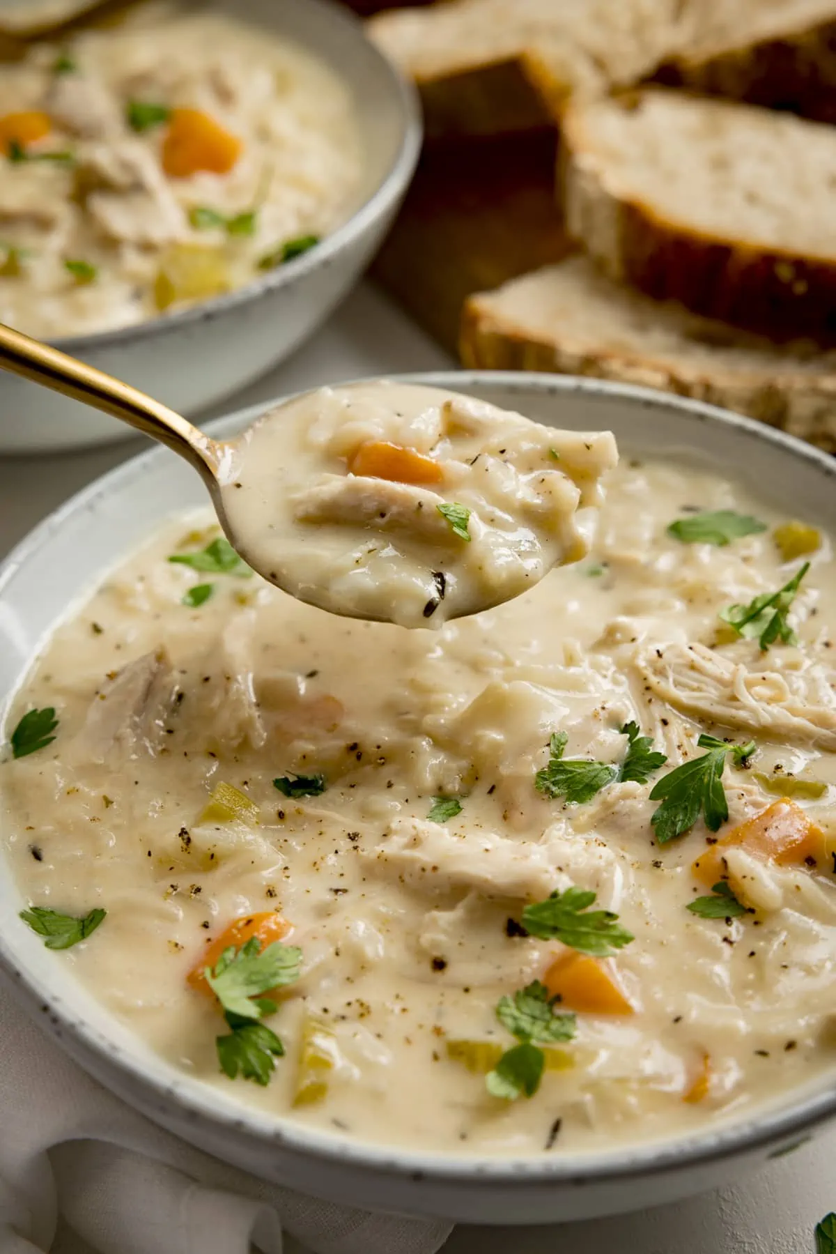 A spoonful being taken from a bowl of chicken and rice soup. There is a further bowl of soup and some bread slices in the background.