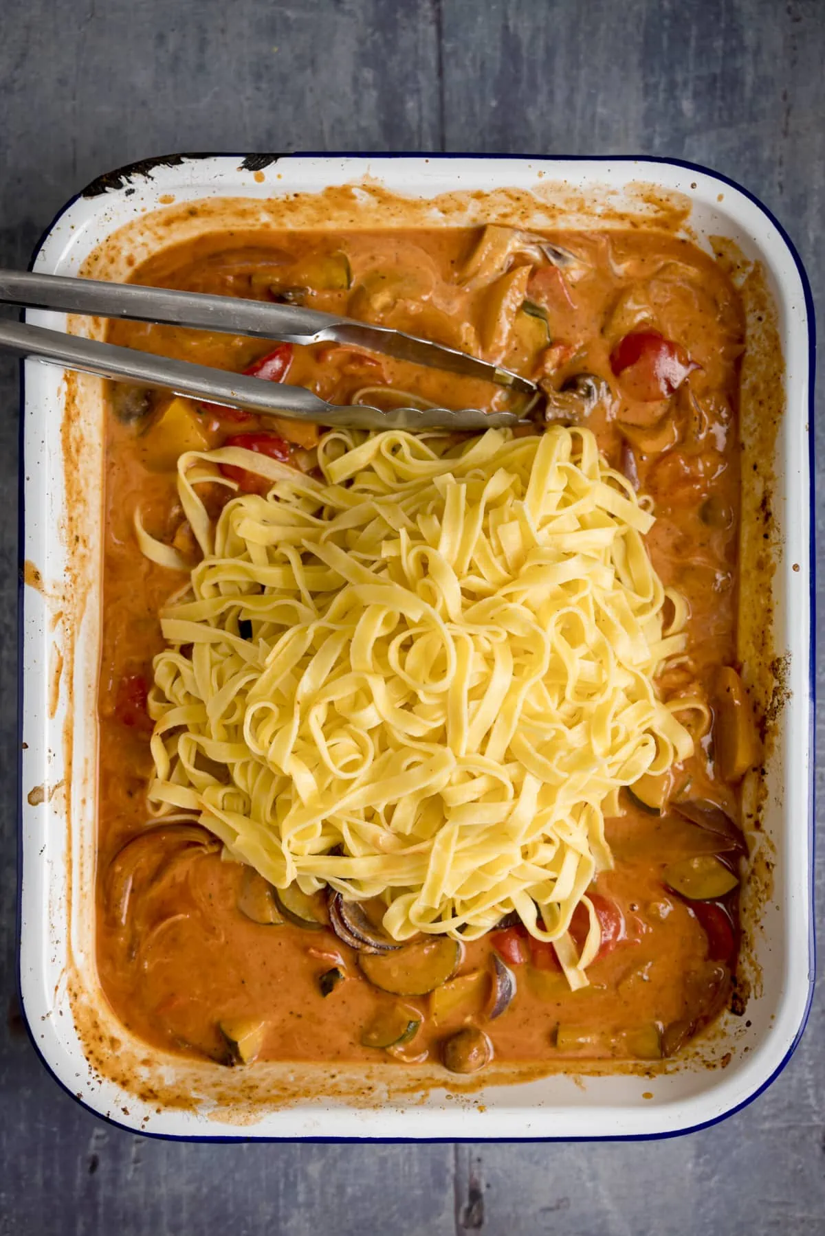 A pile of cooked tagliatelle on top of a roasting tin of roasted vegetable sauce on a blue background. There is a set of tongs in the sauce.