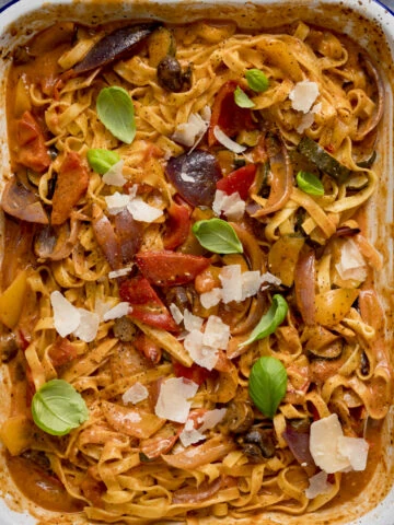 Square image of a white roasting tin filled with tagliatelle in a creamy roasted vegetable sauce on a blue background. The pasta is topped with a few basil leaves and some cheese shavings.