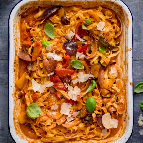 Square image of a white roasting tin filled with tagliatelle in a creamy roasted vegetable sauce on a blue background. The pasta is topped with a few basil leaves and some cheese shavings.