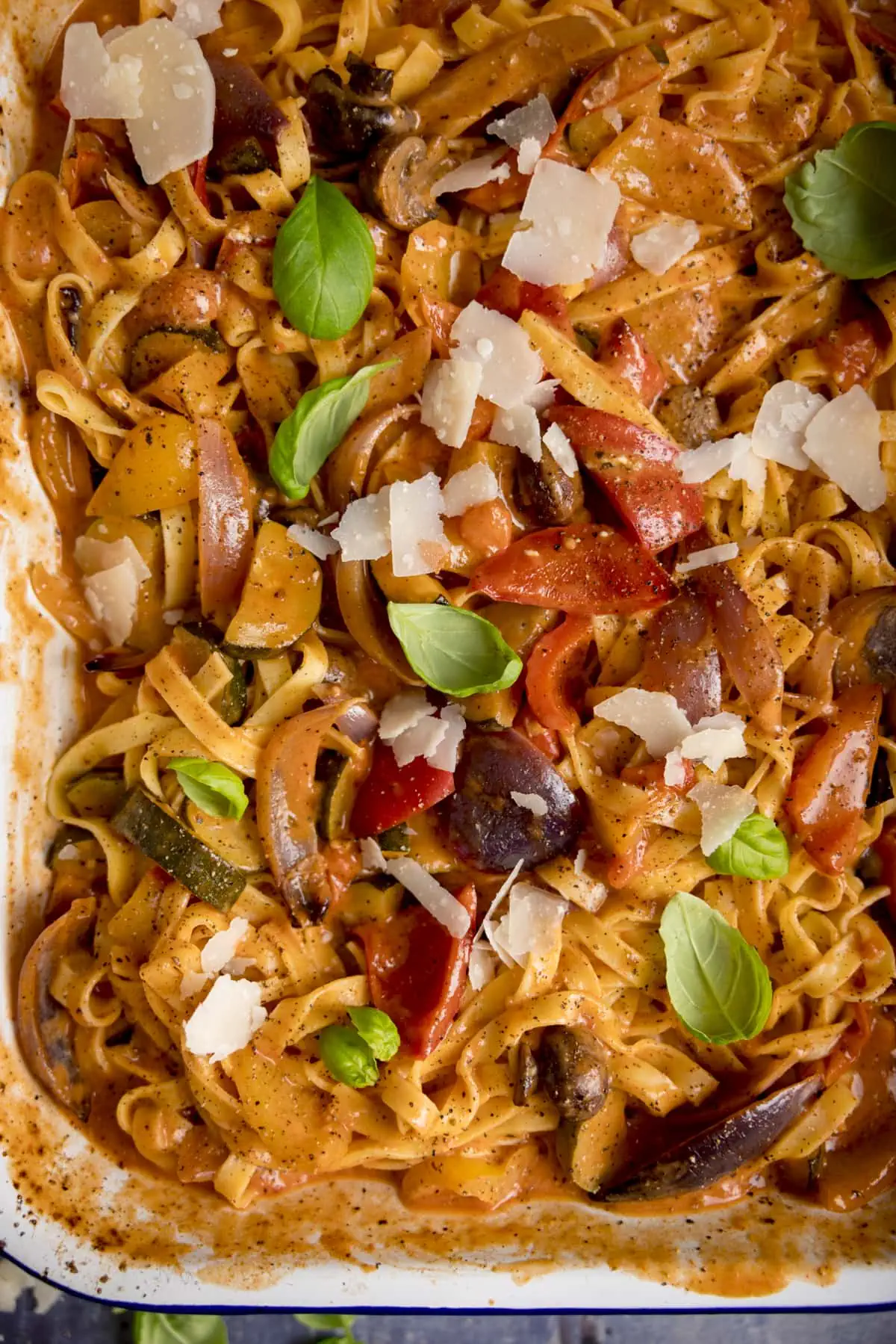 Close up of a white and blue roasting tin filled with tagliatelle in a creamy roasted vegetable sauce. There are basil leaves and cheese shavings sprinkled on top.