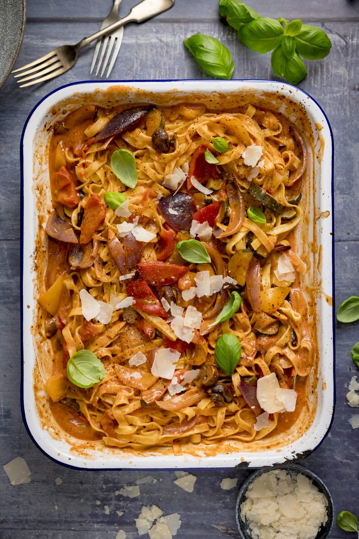 A white roasting tin filled with tagliatelle in a creamy roasted vegetable sauce on a blue background. The pasta is topped with a few basil leaves and some cheese shavings. There are some scattered around the roasting tin too.