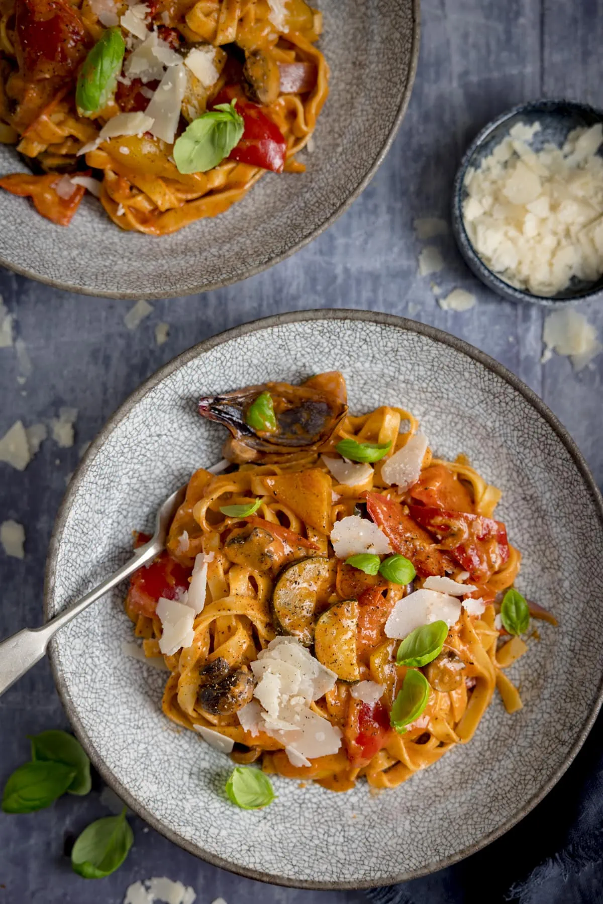 Overhead of a bowl of roasted vegetable tagliatelle on a blue background. There is a further bowl of pasta at the top of the frame and a bowl of shaved cheese in shot.