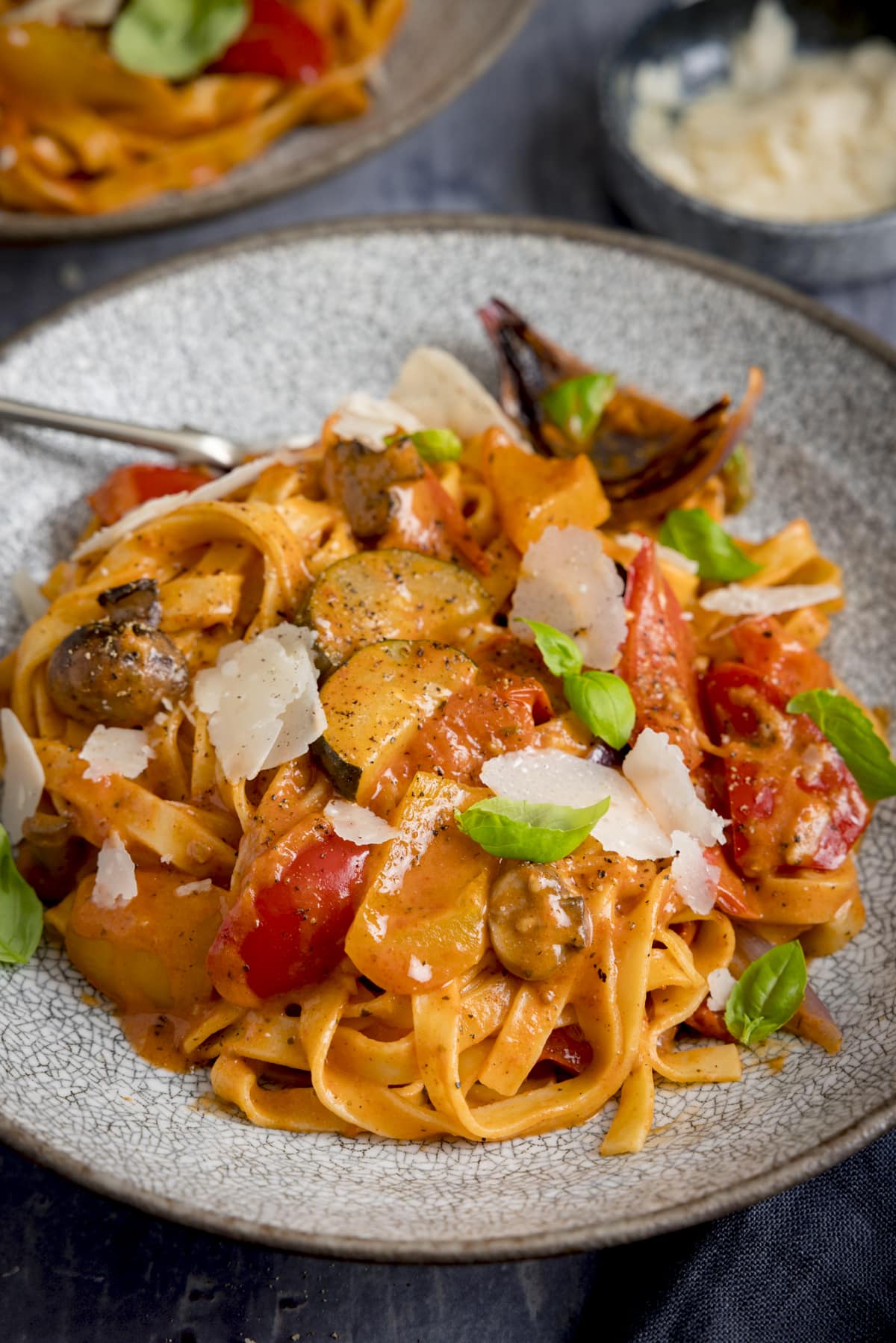 Tall image of a bowl of roasted vegetable tagliatelle on a blue background with ingredients scattered around.