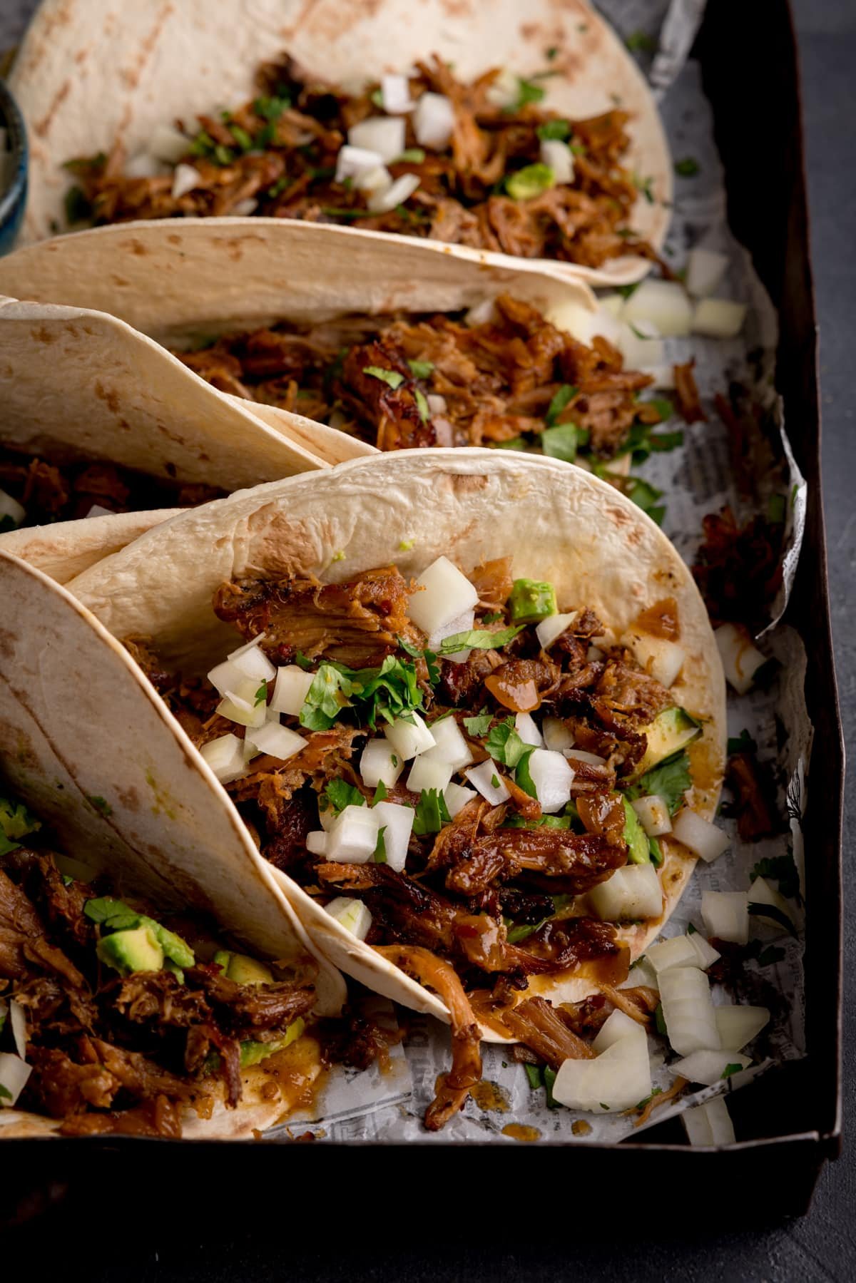 Tall close-up image of pork carnitas tacos on a tray, topped with chopped onion and coriander.
