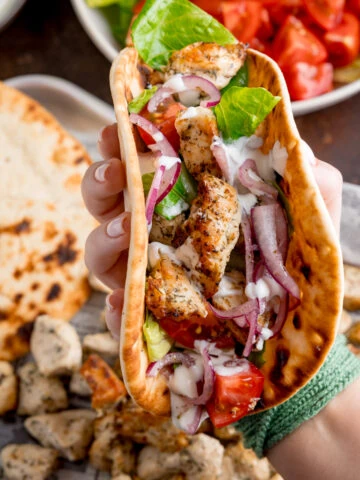 Square overhead shot of air fryer Greek chicken pieces in a flatbread with salad and feta dressing. The wrap is being held by a hand over the top of a tray that has all of the cooked ingredient on it.