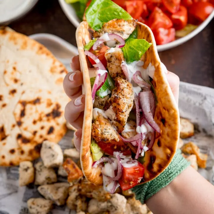 Square overhead shot of air fryer Greek chicken pieces in a flatbread with salad and feta dressing. The wrap is being held by a hand over the top of a tray that has all of the cooked ingredient on it.